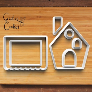 Può includere: Tagliapasta in plastica bianca a forma di casa e cornice rettangolare con bordo smerlato, su una superficie di legno. Il testo "Cactus Cookies" si trova nell'angolo in alto a sinistra.