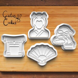 Puede incluir: Cuatro cortadores de galletas de plástico blanco con forma de mujer japonesa, una puerta torii, un árbol bonsái y un abanico. Los cortadores de galletas están sobre una superficie de madera.