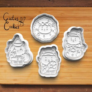 May include: Four white plastic cookie cutters shaped like cats wearing Santa hats. The cutters are on a wooden surface. The text "Cactus Cookies" is written in black ink on the surface.