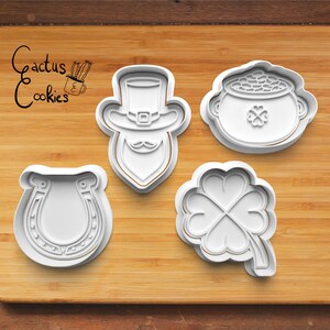 May include: Four white plastic cookie cutters on a wooden surface. The cutters are shaped like a leprechaun, a pot of gold, a four-leaf clover, and a horseshoe.