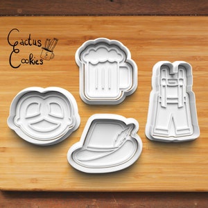 Puede incluir: Cuatro cortadores de galletas de plástico blanco con forma de pretzel, una jarra de cerveza, pantalones de cuero y un sombrero con una pluma. Los cortadores están sobre una superficie de madera.