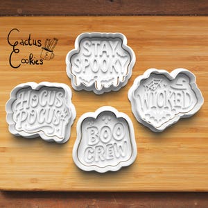 Puede incluir: Cuatro cortadores de galletas blancos con temática de Halloween con las frases "Stay Spooky", "Hocus Pocus", "Wicked" y "Boo Crew". Los cortadores están sobre una superficie de madera, con el logo "Cactus Cookies" en la esquina superior izquierda.