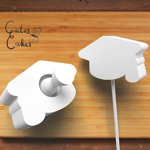 Puede incluir: Dos cortadores de galletas blancos: un gorro de graduación y una forma de mano, sobre un fondo de madera. La imagen incluye el texto "Cactus Cookies".