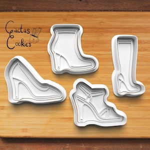 Puede incluir: Cuatro cortadores de galletas de plástico blanco con forma de zapatos y botas de tacón alto. Los cortadores incluyen un tacón de cuña, un tacón de aguja, una sandalia y una bota alta. El logotipo de Cactus Cookies está en la esquina superior izquierda.
