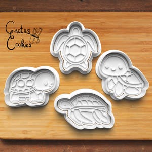 May include: Four white plastic cookie cutters shaped like sea turtles. The cutters are on a wooden surface. The cutters are for making cookies.
