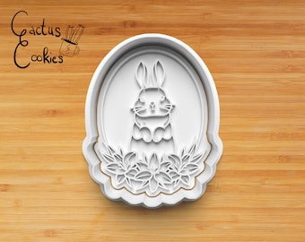 May include: White plastic cookie cutter shaped like an oval with a bunny rabbit inside. The bunny is surrounded by flowers. The cookie cutter is on a wooden surface.