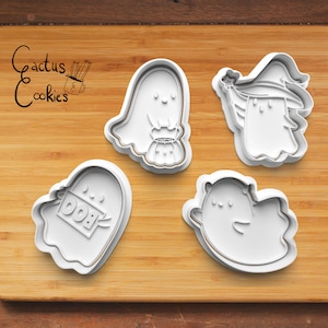 May include: Four white plastic Halloween cookie cutters on a wooden surface. The cutters are shaped like ghosts, with different designs. One ghost has a pumpkin, one has a witch's hat, one has a "BOO" sign, and one has a simple ghost shape.