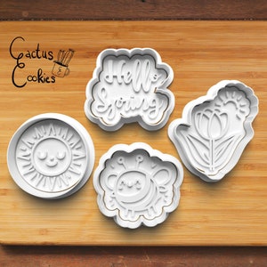May include: Four white plastic cookie cutters on a wooden surface. The cutters are shaped like a sun, a bee, a tulip, and the words "Hello Spring".