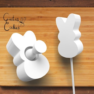 May include: Two white plastic bunny-shaped cookie cutters on a wooden surface. The cutter on the left is a flat shape, while the cutter on the right is a 3D shape with a stick attached.