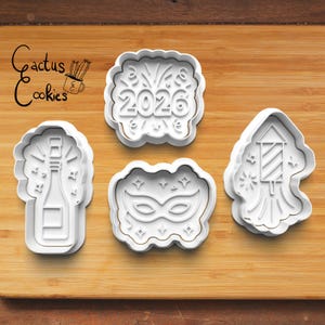 May include: Four white plastic cookie cutters on a wooden surface. The cutters include a champagne bottle, a mask, a firework, and a 2026 design. The Cactus Cookies logo is in the upper left corner.