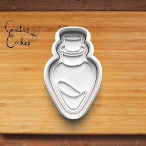 Peut inclure: Un emporte-pièce en plastique blanc en forme de flacon de potion. Le découpoir a une base en forme de cœur et un dessus cylindrique avec un petit bouchon. Les mots "Cactus Cookies" sont écrits en cursive à gauche.