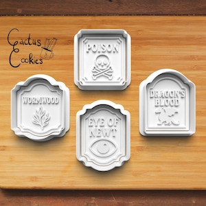May include: Four white cookie cutters with gold trim, arranged on a wooden surface. The cutters are labeled "Poison," "Wormwood," "Eye of Newt," and "Dragon's Blood." The "Poison" cutter features a skull and crossbones.