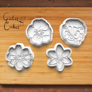 Puede incluir: Cuatro cortadores de galletas de plástico blanco con forma de flores. Los cortadores están sobre una superficie de madera. El texto "Cactus Cookies" es visible en la esquina superior izquierda de la imagen.
