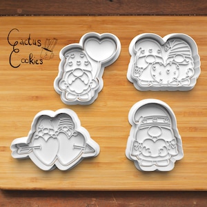 Puede incluir: Cuatro cortadores de galletas de plástico blanco con forma de gnomos que sostienen corazones. Los gnomos llevan sombreros y tienen barbas. Un gnomo sostiene un globo en forma de corazón. El texto "Cactus Cookies" es visible en la esquina superior izquierda de la imagen.
