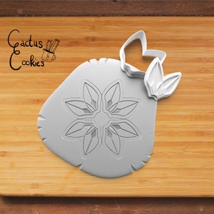 May include: A gray plastic cookie cutter with a floral design and a separate leaf-shaped piece. The cutter has the text "Cactus Cookies" in the upper left corner. The cutter is on a wooden surface.