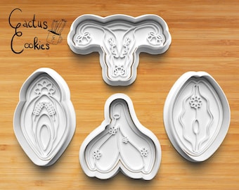 May include: Set of three white plastic cookie cutters shaped like female reproductive organs. The cutters are on a wooden surface.