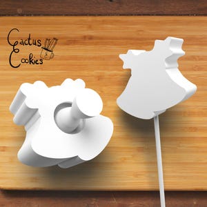 以下が含まれることがあります： 白いドレス型のクッキーカッターが2つ。1つは持ち手付き、もう1つは棒付き。木製の表面に置かれています。「Cactus Cookies」の文字が左上にあります。