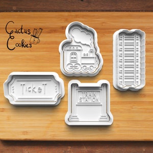 Puede incluir: Cuatro cortadores de galletas de plástico blanco con forma de tren, vías de tren, un letrero de estación de tren y un billete. Los cortadores están sobre una superficie de madera.
