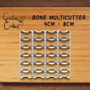 May include: A white plastic bone multicutter featuring a grid of twenty-four connected bone-shaped cookie cutters. The text "Cactus Cookies" and "BONE MULTICUTTER 4CM - 8CM" is printed above the cutter. The tool is designed for cutting multiple shapes.