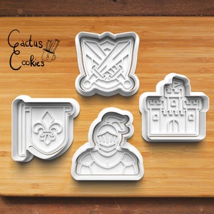 May include: Set of four white plastic cookie cutters shaped like a knight, a castle, a shield with swords, and a banner with a fleur-de-lis. The cookie cutters are on a wooden surface.