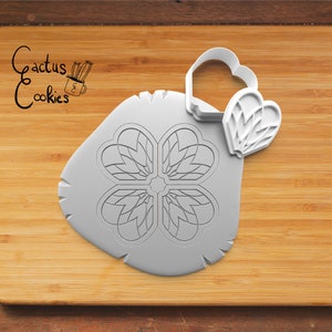 May include: A white cookie cutter with a four-petal floral design, next to a heart-shaped cutter. The design is pressed into a piece of gray dough. The image is on a wooden surface. The text "Cactus Cookies" is in the upper left corner.