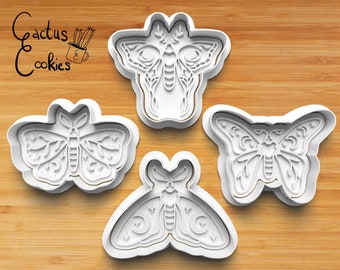 May include: Four white plastic cookie cutters shaped like moths with intricate designs. The cutters are on a wooden surface.