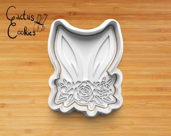 May include: White plastic cookie cutter shaped like a bunny rabbit with a floral crown. The cutter is on a wooden surface.