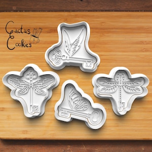 May include: Four white plastic cookie cutters shaped like a dragonfly, a butterfly, a key with wings, and a key with a dragonfly. The cutters are on a wooden surface. The text "Cactus Cookies" is in the top left corner of the image.