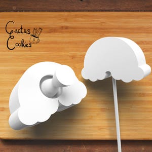 May include: Two white cookie cutters in the shape of a cloud and a cloud with a stick. The cookie cutters are on a wooden surface. The text "Cactus Cookies" is in the upper left corner.