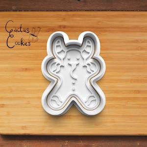 May include: White plastic gingerbread man cookie cutter with a smiling face and raised arms. The cutter is on a wooden surface.