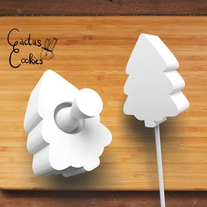 Puede incluir: Dos cortadores de galletas de árbol de Navidad de plástico blanco sobre una superficie de madera. El cortador más grande tiene un mango y el cortador más pequeño es plano.