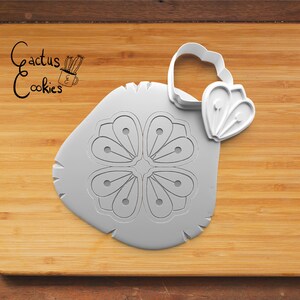 May include: A gray plastic cookie cutter in the shape of a four-petal flower with a handle. The cutter is on a wooden surface. The text "Cactus Cookies" is in the upper left corner.