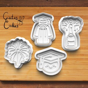 May include: Four white plastic cookie cutters on a wooden surface. The cutters are shaped like a graduation cap, a graduation gown, a bottle of champagne with a firework design, and a firework.