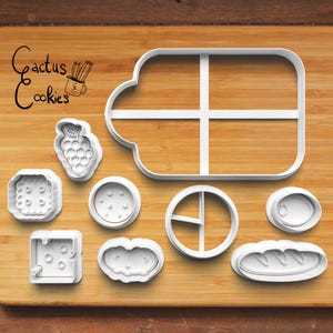 May include: A collection of white plastic cookie cutters in various shapes, including a grape cluster, a square with holes, a circle with dots, a circle with a line, an oval, and a loaf of bread. The image also includes a large bus-shaped cutter and the text "Cactus Cookies".