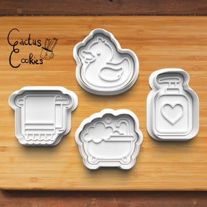 May include: Four white plastic cookie cutters in the shape of a rubber duck, a towel on a rack, a bathtub with bubbles, and a soap dispenser with a heart. The text "Cactus Cookies" is in the upper left corner.