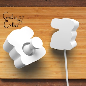 May include: Two white plastic molds for making bunny-shaped cookies. One mold is a flat bunny shape, the other is a bunny with a stick attached for making a lollipop.