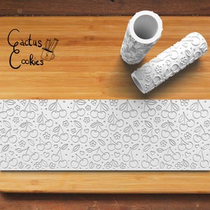 May include: White rolling pins with textured patterns of cherries, flowers, and hearts. A white rectangular sheet with the same cherry pattern is also visible. The image includes the text "Cactus Cookies".