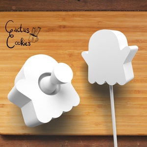 May include: White ghost-shaped cookie cutters on a wooden surface. One cutter has a handle, and the other is on a stick. The text "Cactus Cookies" is in the upper left corner.