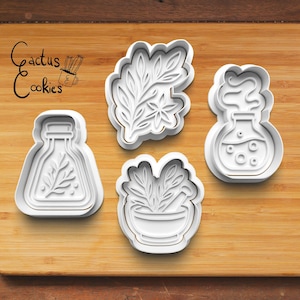 Puede incluir: Cuatro cortadores de galletas de plástico blanco con forma de matraz, ramita de hojas, mortero y maja, y matraz con vapor. El texto "Cactus Cookies" está en la esquina superior izquierda. Los cortadores están sobre una superficie de madera.