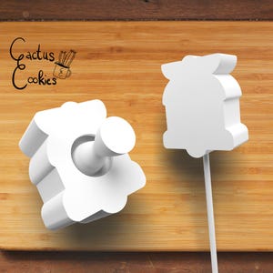以下が含まれることがあります： 白い花の形をしたクッキーカッターが2つ。1つは持ち手付き。木製の表面に置かれています。「Cactus Cookies」の文字が左上にあります。焼き菓子作りに最適です。