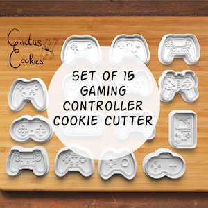 Może przedstawiać: Zestaw 15 białych plastikowych foremek do ciasteczek w kształcie różnych kontrolerów do gier wideo. Formy są ułożone na drewnianej powierzchni. Tekst "SET OF 15 GAMING CONTROLLER COOKIE CUTTER" jest nadrukowany czarnym kolorem na białym kole w środku obrazu.