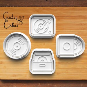 May include: Four white plastic cookie cutters with music-themed designs including a circular vinyl record, a square record player, a rectangular cassette tape, and a storage crate with records. The text Cactus Cookies is printed in black.