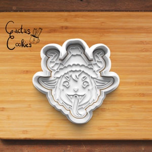 May include: White plastic cookie cutter shaped like a cartoon monster wearing a Santa hat. The monster has horns, a big smile, and a long tongue sticking out.