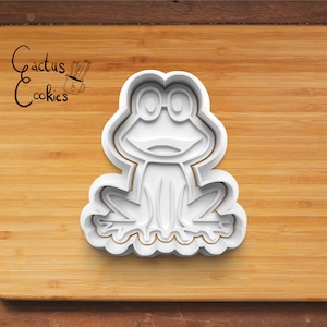 Puede incluir: Cortador de galletas de plástico blanco con forma de rana con ojos grandes y una amplia sonrisa. El cortador está sobre una superficie de madera.