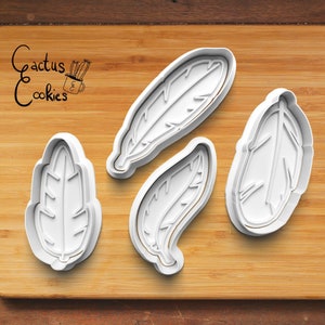 Könnte beinhalten: Drei weiße Plastik-Ausstecher in Form von Federn auf einer Holzoberfläche. Die Ausstecher sind zum Ausstechen von Keksen in Form von Federn.