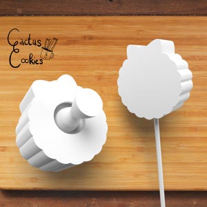 May include: Two white, flower-shaped cookie cutters on a wooden surface. One cutter has a handle, and the other is on a stick. The text "Cactus Cookies" is in the upper left corner.