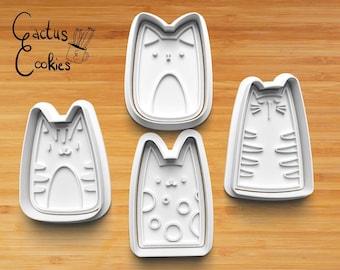 Puede incluir: Cuatro cortadores de galletas de plástico blanco en forma de gato sobre una superficie de madera. Los cortadores presentan diferentes diseños, incluyendo un gato rayado, un gato con lunares y un gato liso.