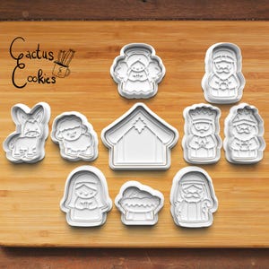 Puede incluir: Un conjunto de cortadores de galletas de pl&aacute;stico blanco con formas variadas, incluyendo un &aacute;ngel, un burro, una oveja, un establo y figuras del nacimiento. Los cortadores est&aacute;n dispuestos sobre una superficie de madera, con el texto "Cactus Cookies".