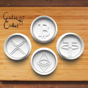 May include: Four white cookie cutters shaped like cryptocurrency logos: Bitcoin, Ethereum, Tether, and XRP. The cutters are on a wooden surface with the text "Cactus Cookies" in black.
