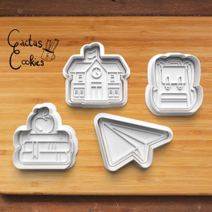 Puede incluir: Cuatro cortadores de galletas de plástico blanco con forma de edificio escolar, mochila, pila de libros con una manzana en la parte superior y avión de papel.